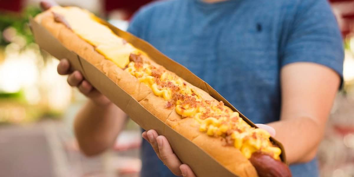 A person in a blue shirt holds one of the Best Disney Hot Dogs—a very long hot dog topped with melted cheese and bacon bits, served in a cardboard tray.