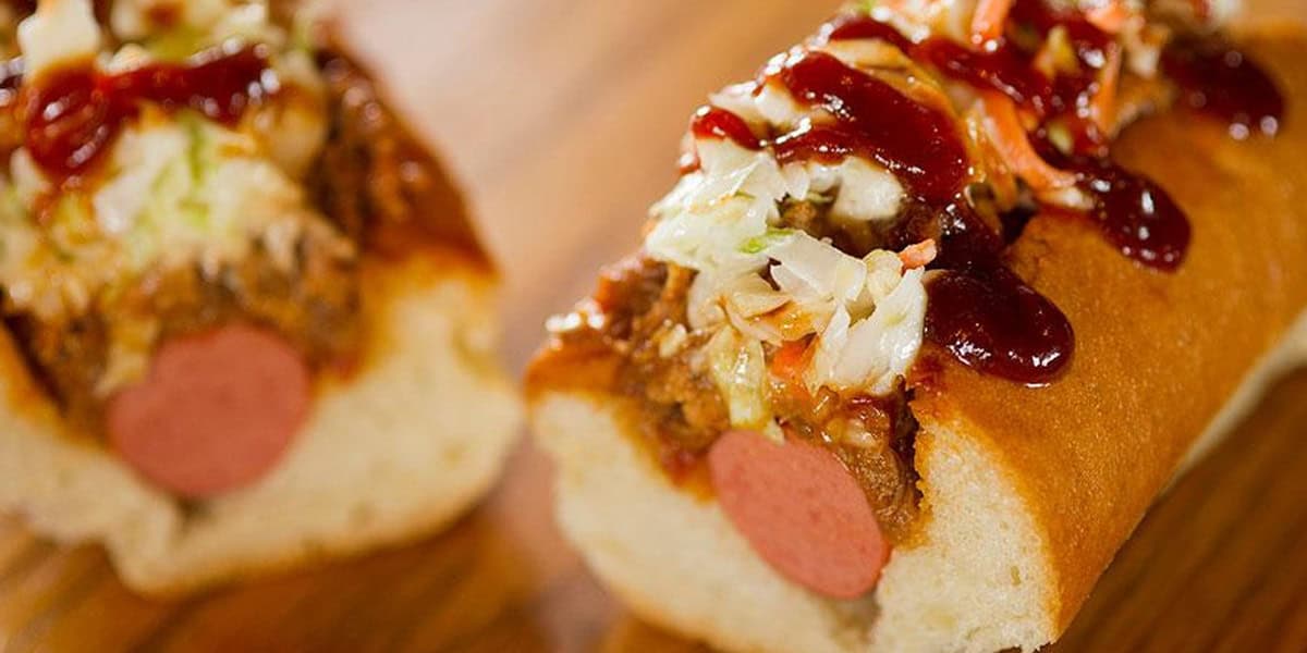 A close-up of one of the Best Disney Hot Dogs in a bun, topped with pulled pork, coleslaw, and barbecue sauce, with another half of the hot dog in the background on a wooden surface.
