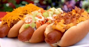 Three hot dogs on a white plate, each topped differently—one with shredded cheddar cheese, one with diced celery and sauce, and one with macaroni and cheese plus bacon bits—just like some of the best Disney hot dogs you'd find in the parks.