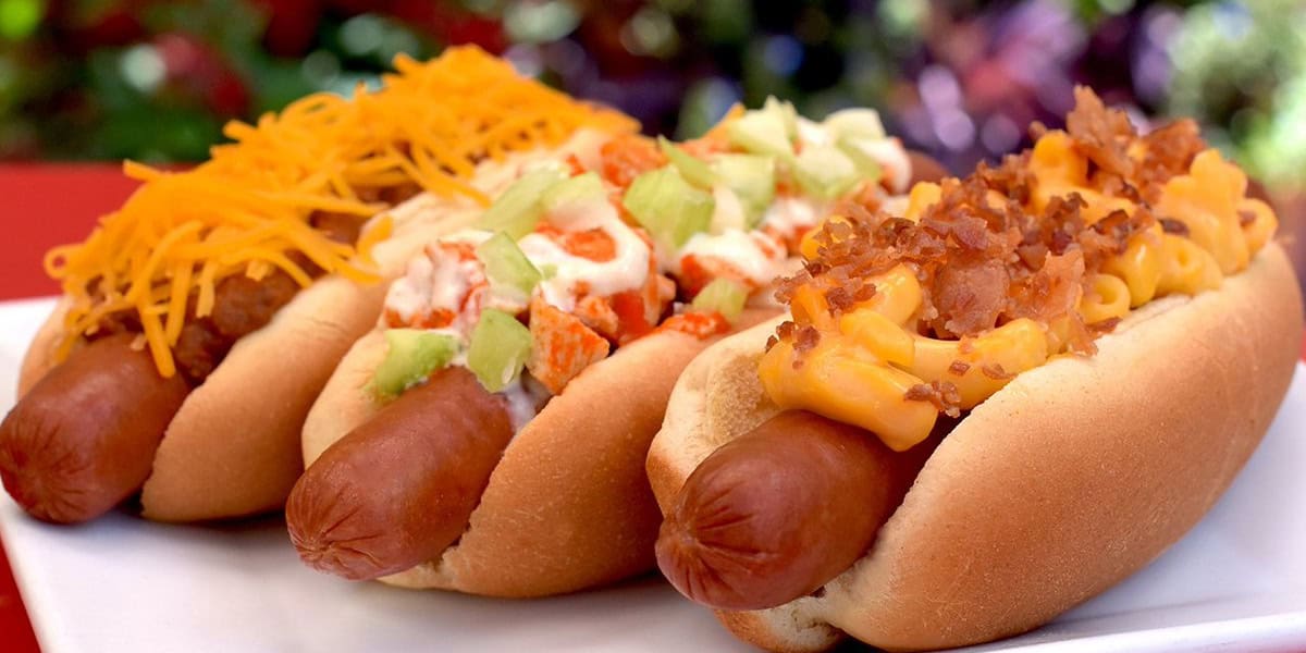 Three hot dogs on a white plate, each topped differently—one with shredded cheddar cheese, one with diced celery and sauce, and one with macaroni and cheese plus bacon bits—just like some of the best Disney hot dogs you'd find in the parks.