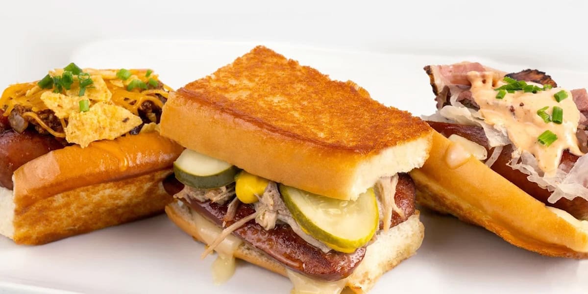 Three gourmet sandwiches on a white plate: one with pickles and pulled pork, another topped with cheese and chips, and a third with sauerkraut, meat, and a creamy sauce—each one rivaling the creativity of the Best Disney Hot Dogs.