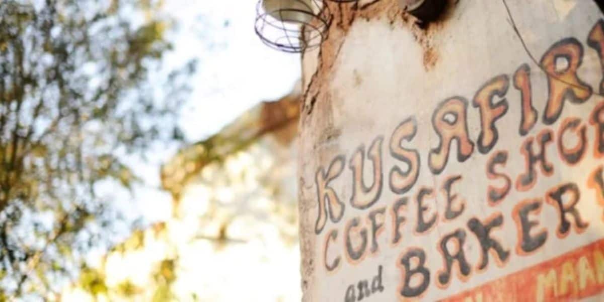 A rustic outdoor sign for "Kusafiri Coffee Shop and Bakery" is mounted on a wall, partially obscured by tree branches. The sign features vintage-style lettering and a weathered appearance.