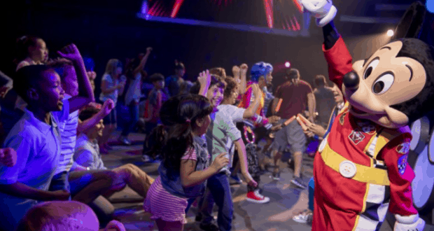 Kids dance and cheer around Mickey Mouse at the Disney Junior Dance Party, surrounded by vibrant, colorful lights indoors.