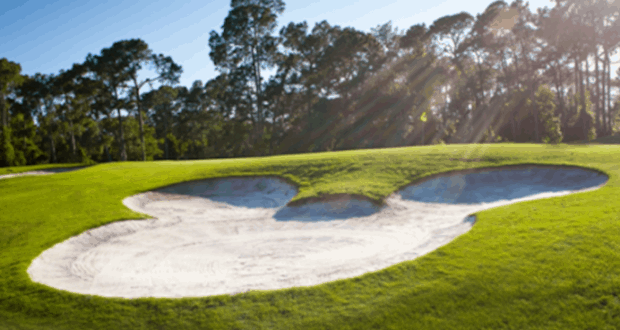 A Mickey Mouse-shaped sand bunker on a sunlit Disney World golf course, surrounded by lush green grass and trees.