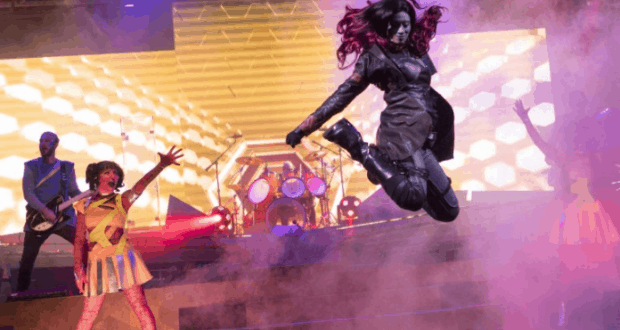 A Guardians of the Galaxy performer leaps mid-air at Awesome Mix Live, with vibrant lights, live band, and backup dancers on stage.
