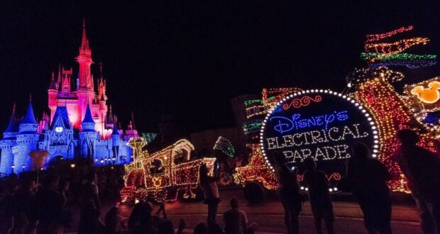 A vibrant night scene at Disney's Electrical Parade, featuring an illuminated float with a sign reading "Disney's Electrical Parade." A castle glowing with colorful lights stands in the background, as silhouettes of people watch the spectacle.