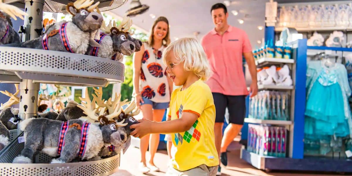 A young child in a yellow shirt excitedly reaches for a plush reindeer in a store. Two adults smile in the background. The store is filled with stuffed toys and costumes, creating a colorful and cheerful atmosphere.