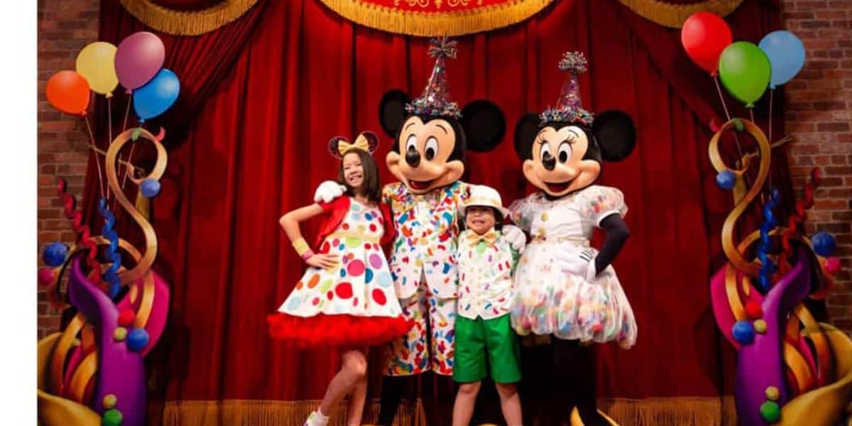 Two children in colorful outfits stand between two costumed characters resembling mice in festive clothes. They are all smiling on a stage decorated with balloons and a red curtain.