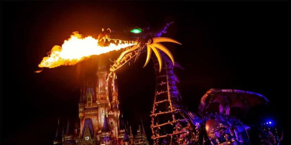 A large, mechanical dragon breathes fire in front of an illuminated castle during a nighttime parade. The dragon has glowing green eyes and a spiked metallic body, with a dark sky in the background.