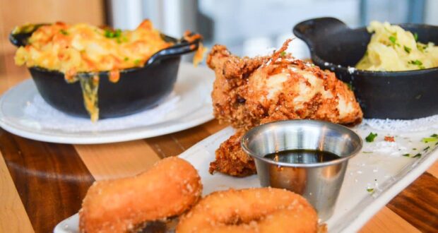 A close-up of comfort food featuring fried chicken, mini donuts, syrup, mashed potatoes, and cheesy macaroni and cheese—just like the Best Table Service Meals at Disney—served on white plates and bowls atop a wooden table.