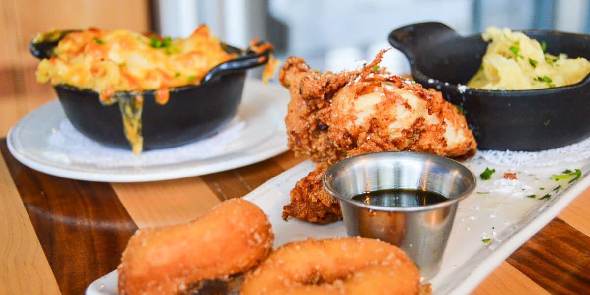 A close-up of comfort food featuring fried chicken, mini donuts, syrup, mashed potatoes, and cheesy macaroni and cheese—just like the Best Table Service Meals at Disney—served on white plates and bowls atop a wooden table.