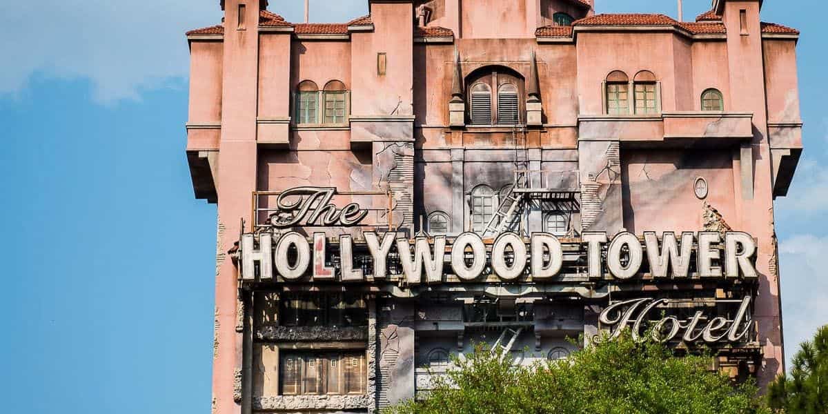 The image shows the Hollywood Tower Hotel, a tall, intricately designed building with a weathered facade and an ornate sign. The structure has a pinkish hue, and the sky is blue with a few clouds in the background. Trees partially obscure the view.