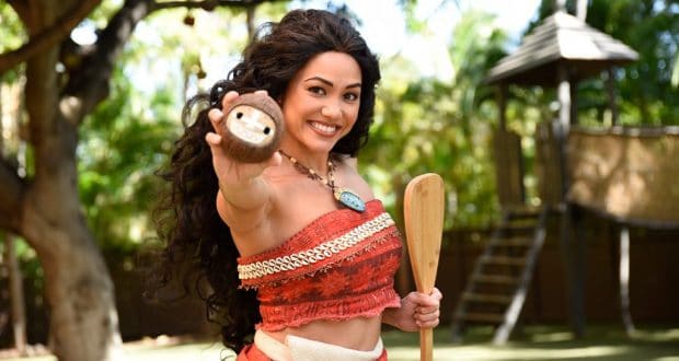 Moana in her signature red outfit with long curly hair smiles, holding a paddle and prop during a Walt Disney World meet and greet.
