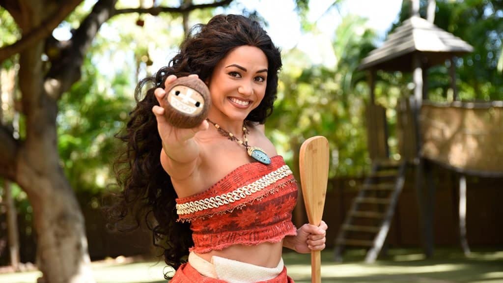 Moana in her signature red outfit with long curly hair smiles, holding a paddle and prop during a Walt Disney World meet and greet.