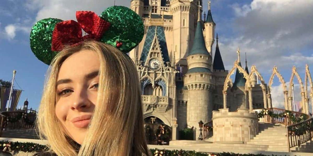 A woman with long blonde hair wears green and red Mickey Mouse ears. She is smiling in front of a castle at a theme park, with blue sky and clouds in the background.