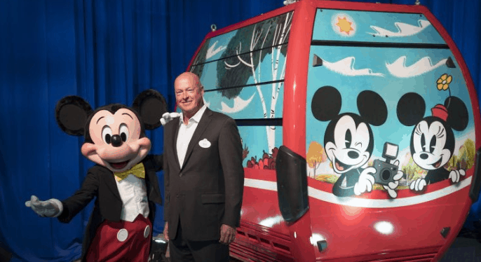 Mickey Mouse greets a guest next to a Disney Skyliner gondola at Walt Disney World, featuring cheerful Mickey and Minnie designs.
