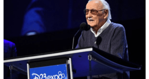 An elderly man wearing sunglasses and a sweater speaks at a podium with a sign that reads "D23 Expo: The Ultimate Disney Fan Event." The background is dark and the scene is brightly lit.