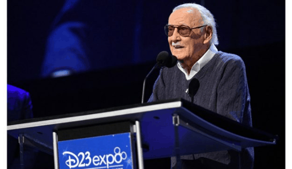 An elderly man wearing sunglasses and a sweater speaks at a podium with a sign that reads "D23 Expo: The Ultimate Disney Fan Event." The background is dark and the scene is brightly lit.