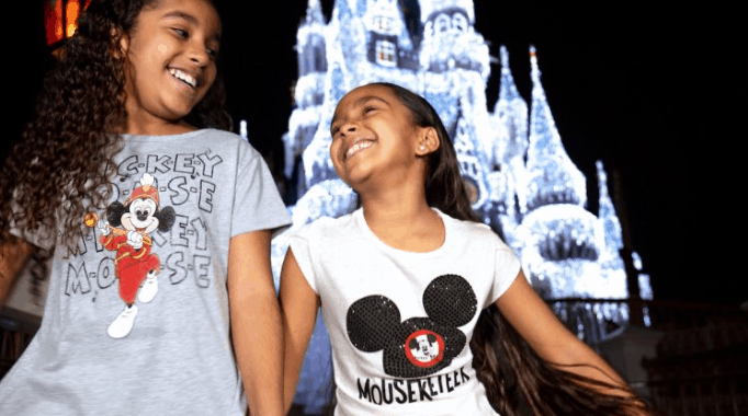 Two girls in Mickey Mouse shirts pose by a glowing Cinderella Castle at night, celebrating the magic of Disney World together.