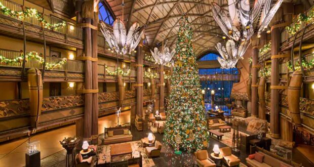 A grand hotel lobby decorated for the holidays with a towering Christmas tree adorned with lights and ornaments. Festive garlands wrap around the balconies and large, unique chandeliers hang from the ceiling. Comfortable seating areas surround the tree.