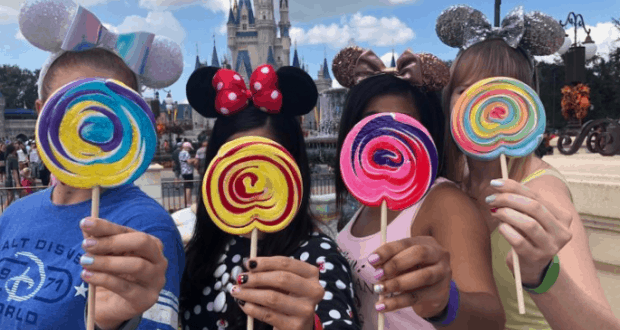 Friends in Mickey ears pose at Disney World, faces hidden by rainbow lollipops; Disney nail art adds flair to the fun atmosphere.