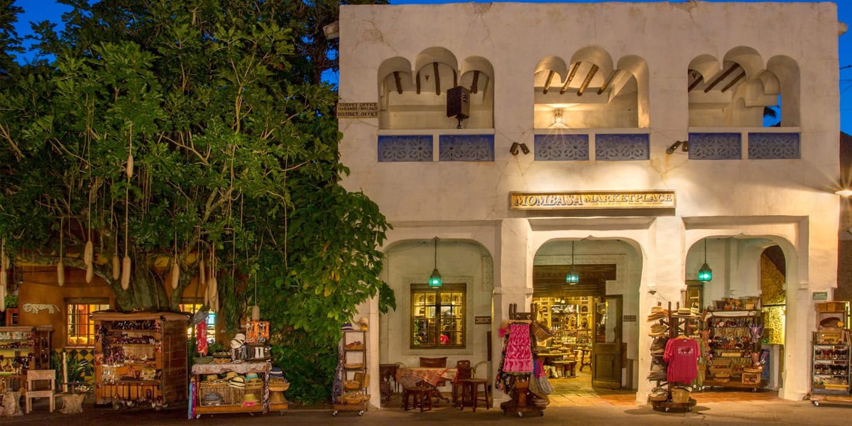 A white stucco building with arched windows and wooden beams houses the Mombasa Marketplace in Africa at Animal Kingdom, featuring outdoor displays of colorful souvenirs, clothing, and crafts, surrounded by lush greenery at dusk.