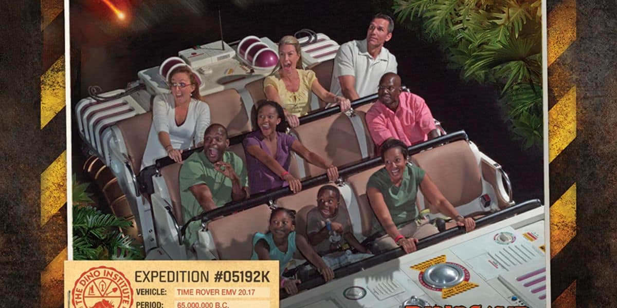 A group of people, including children and adults, are seated in a theme park ride vehicle, experiencing a thrilling moment. They appear excited and surprised as they descend on the ride. The seating resembles a safari expedition vehicle.