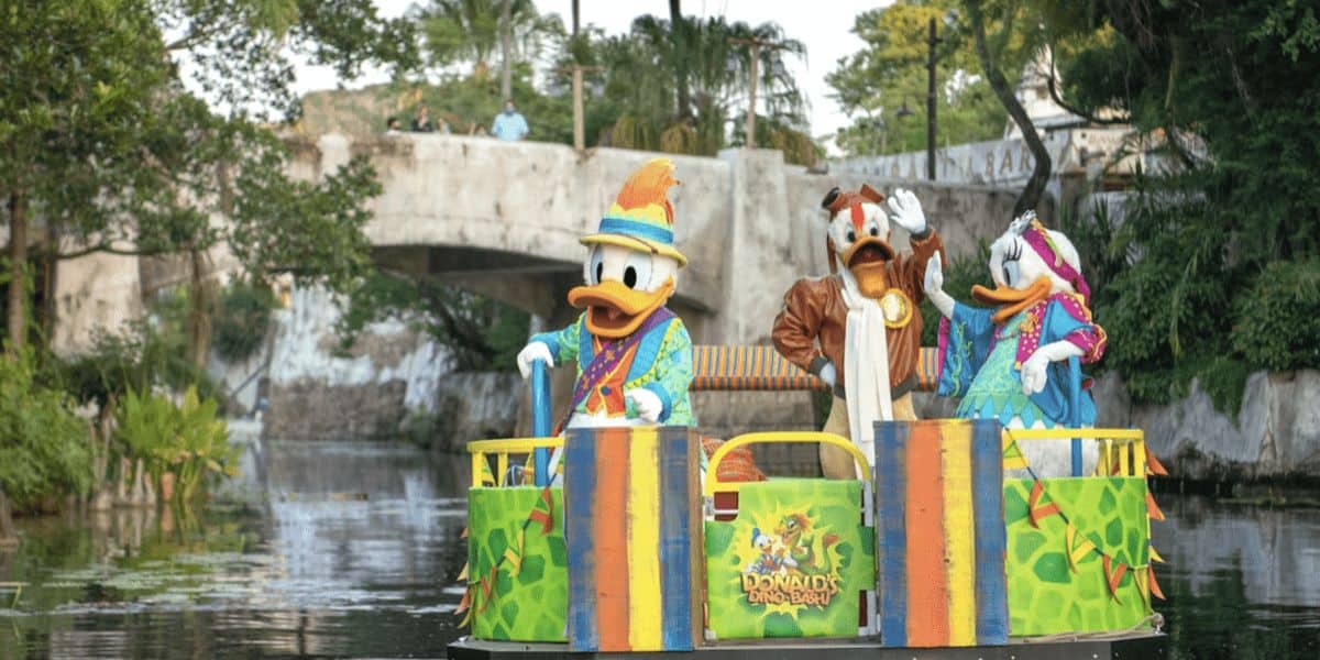 Three cartoon characters on a colorful float decorated with jungle-themed elements and a "Donald's Dino Bash" sign, cruising on a river. There's lush greenery and a stone bridge in the background.