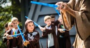 Children dressed as Jedi wielding blue lightsabers are being guided by an adult in a similar robe. The group is outdoors, standing in line, with focused expressions as they practice. A young boy in the foreground wears a robe and a Mickey Mouse shirt.