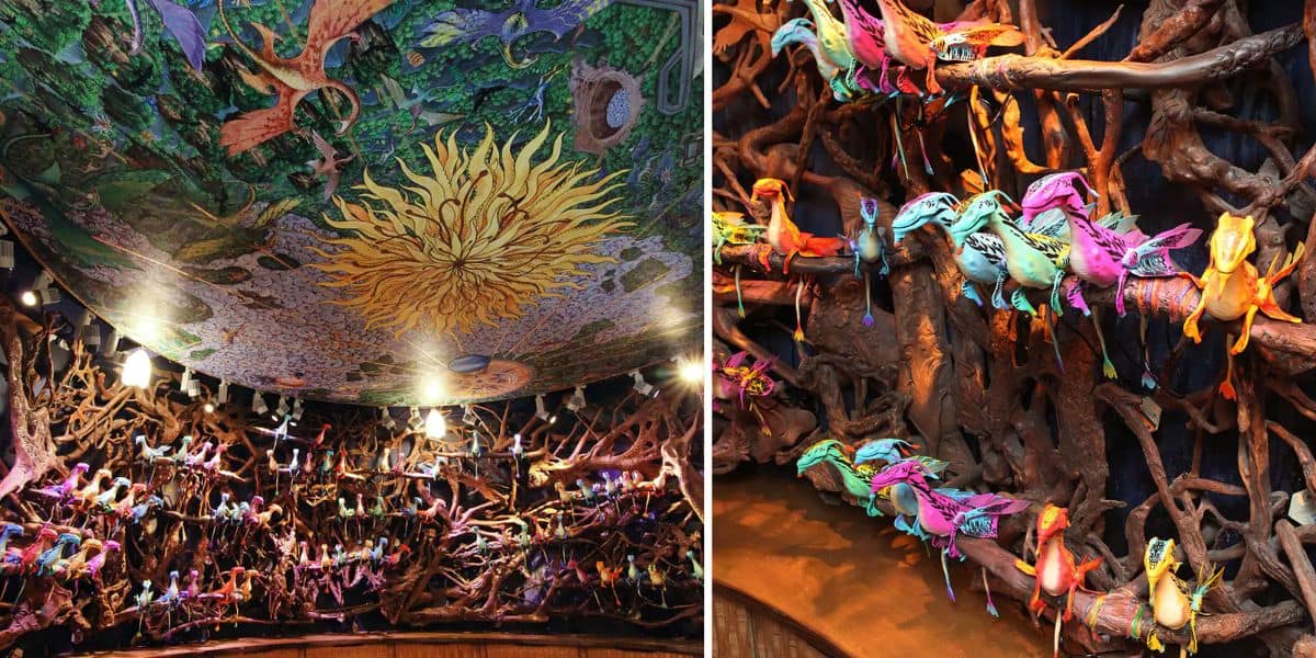 Colorful tropical birds perch on twisted branches inside a vibrant, indoor sanctuary. The ceiling features an intricate mural with a swirling, sun-like design and lush, green foliage, creating an immersive, naturalistic environment.