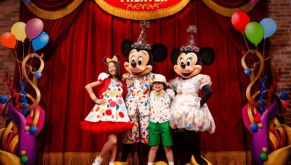 Mickey and Minnie Mouse in festive attire join two kids on a vibrant stage, surrounded by balloons at Disney World’s celebration.