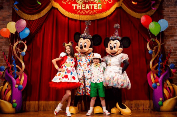 Mickey and Minnie Mouse in festive attire join two kids on a vibrant stage, surrounded by balloons at Disney World’s celebration.