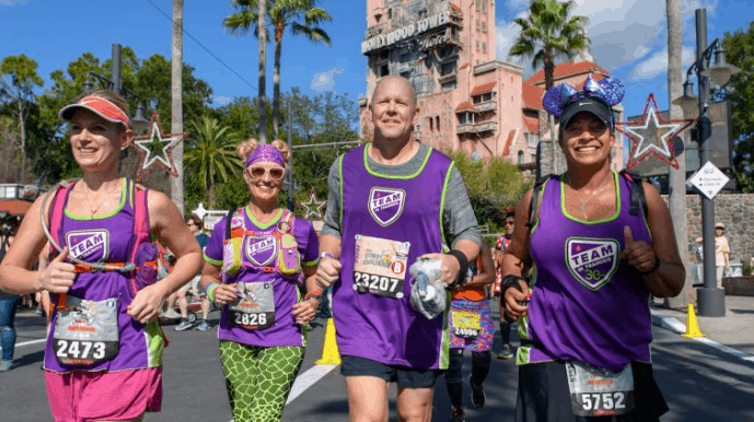 Marathon runners in purple jerseys race past palm trees and the iconic Disney World tower during a magical runDisney event.