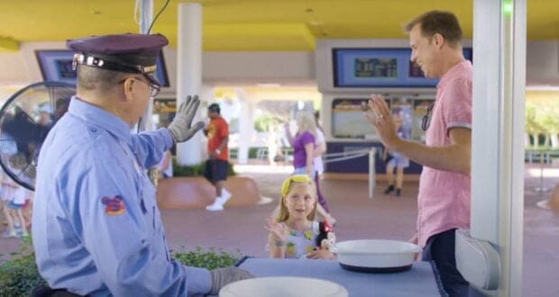 A Disney World security guard in uniform waves to a smiling young girl and an adult man, both raising their hands in greeting, at an outdoor checkpoint.