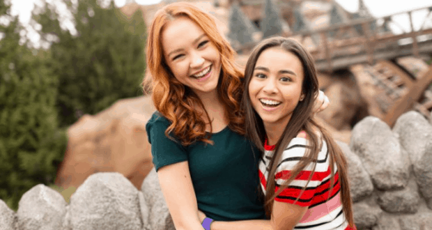 Two friends embrace by a stone fence at Disney’s Hollywood Studios, channeling Kim Possible with trees and classic park scenery behind.