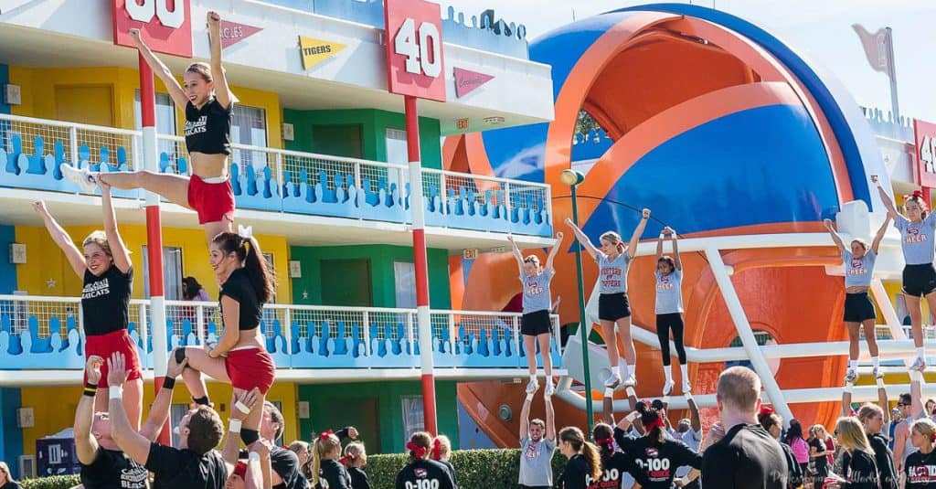 Cheerleaders wow guests with acrobatic stunts in front of Disney’s vibrant All-Star Resorts, beside a giant blue-orange sports ball.