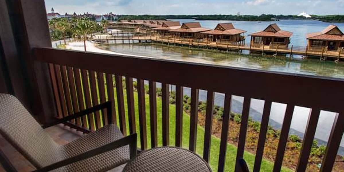 View from a balcony with two chairs, overlooking waterfront bungalows on stilts above a calm lagoon at Disney’s Polynesian Village Resort, surrounded by lush landscaping and distant trees under a partly cloudy sky.