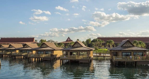Several wooden overwater bungalows with porches sit above calm water at Disney’s Polynesian Village Resort, backed by lush landscaping and red-roofed buildings under a blue sky with scattered clouds.
