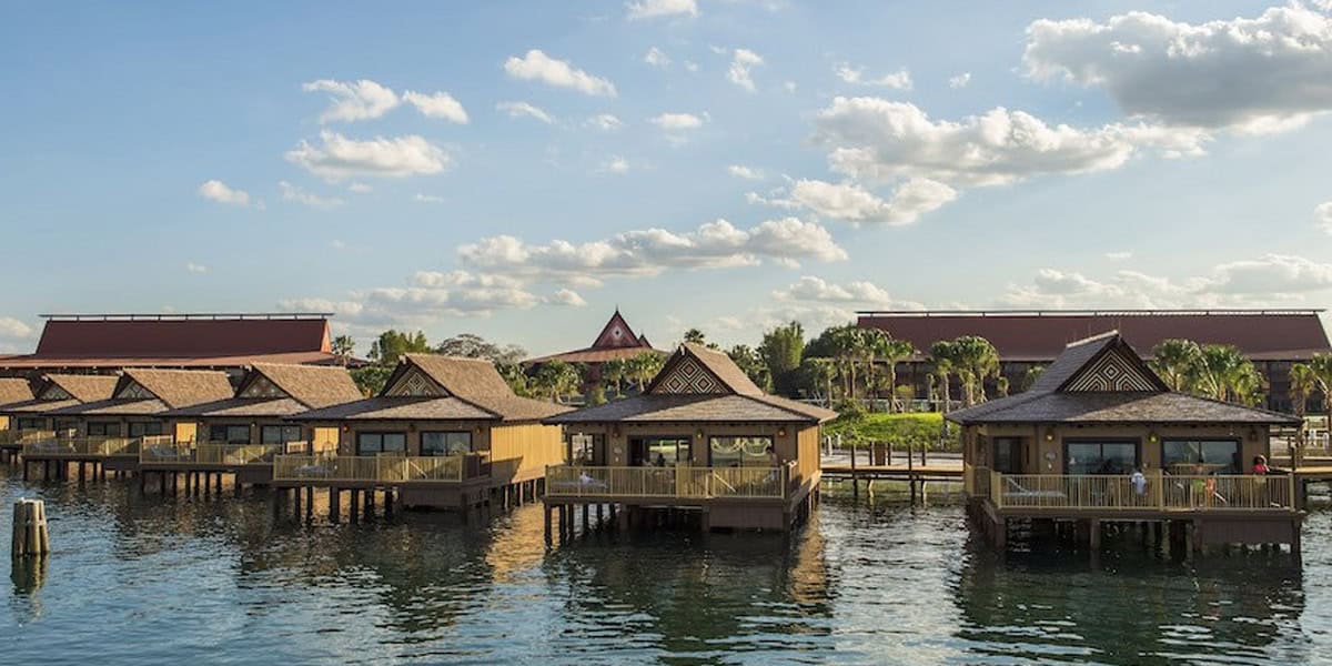 Several wooden overwater bungalows with porches sit above calm water at Disney’s Polynesian Village Resort, backed by lush landscaping and red-roofed buildings under a blue sky with scattered clouds.