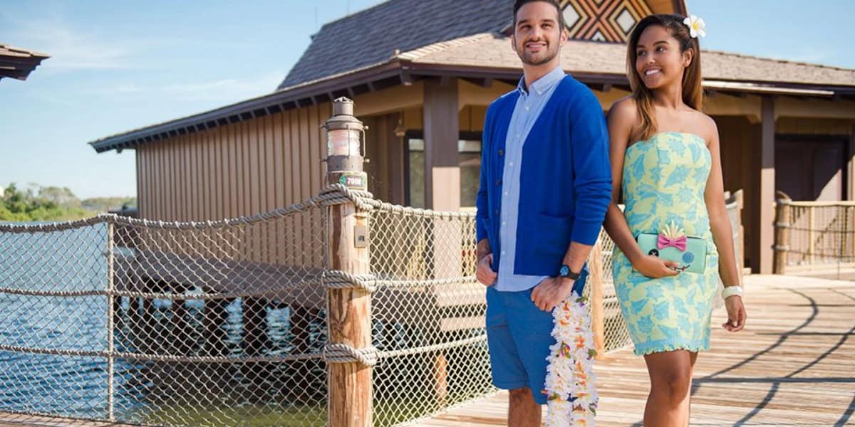 A man and woman stand on a wooden walkway by the water at Disney’s Polynesian Village Resort, near a thatched-roof building. The man wears a blue cardigan, blue shorts, and holds a lei. The woman wears a light blue and yellow dress with a small clutch.