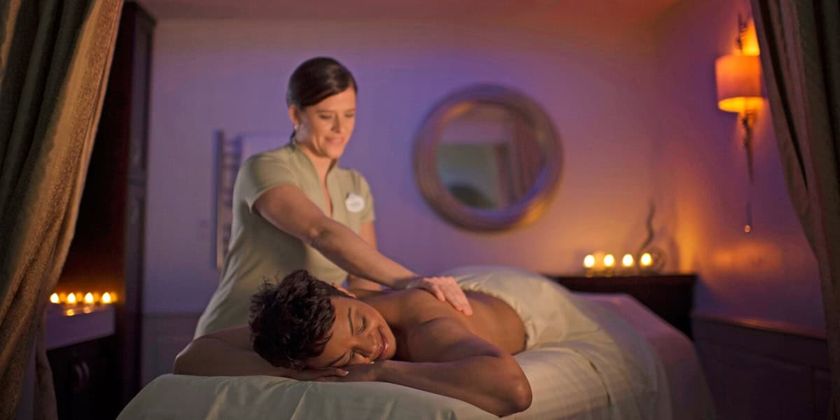A massage therapist gives a relaxing back massage to a person lying face down on a table in a dimly lit spa room with soft lighting and candles, offering the perfect escape at Disney Resorts for Adults.