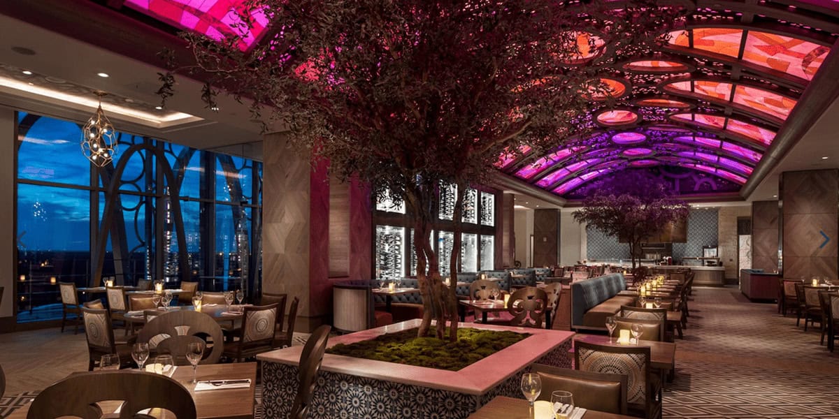 Elegant restaurant interior with large windows, tree planters, and a vibrant arched ceiling of purple and pink stained-glass panels—perfect for guests seeking Disney Resorts for Adults in a warm, inviting atmosphere set for dining.