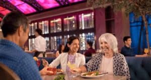 Three people sit at a restaurant table, smiling and enjoying a meal together. The lively atmosphere evokes the vibrant dining experiences found at Disney Resorts for Adults, with colorful lighting and attentive service all around.