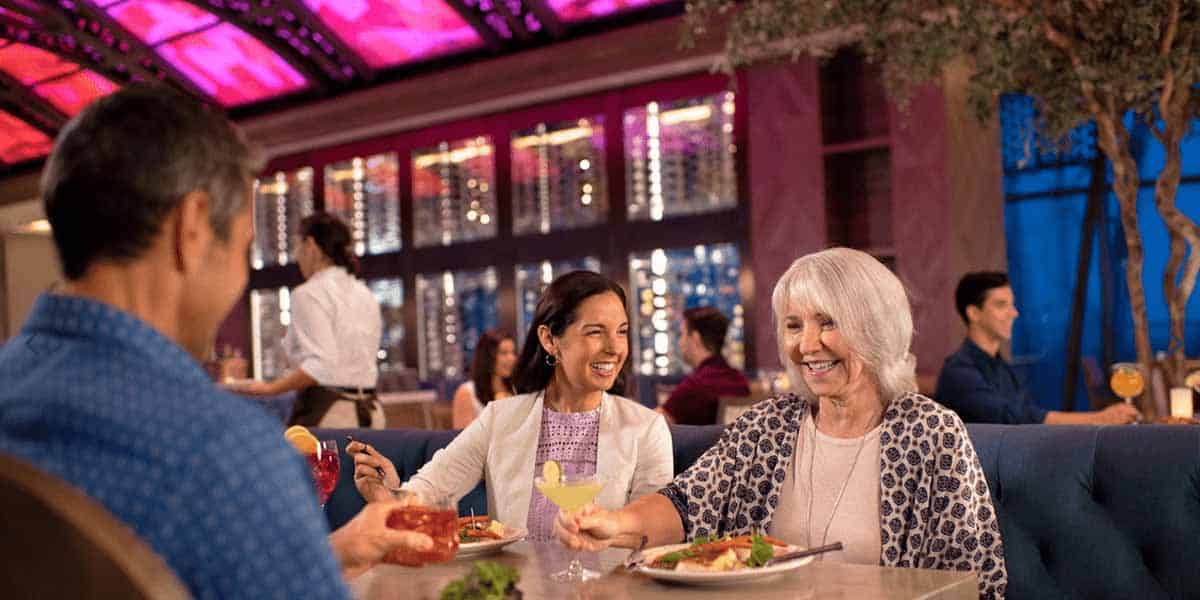 Three people sit at a restaurant table, smiling and enjoying a meal together. The lively atmosphere evokes the vibrant dining experiences found at Disney Resorts for Adults, with colorful lighting and attentive service all around.
