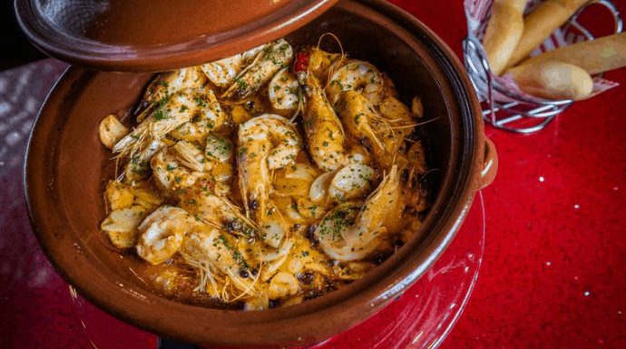 Shrimp and herbs sizzle in a clay pot beside breadsticks on a red table at Jaleo, Disney Springs’ lively dining hotspot.