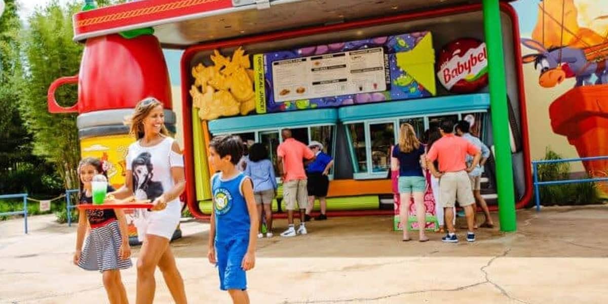 A woman and two children walk past a colorful outdoor food stand shaped like lunchbox items, including a giant Babybel cheese and ketchup bottle—one of the Best Photo Spots at Disney—while others order at the window.