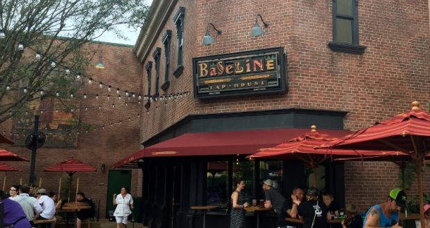 Guests relax at outdoor tables under red umbrellas at Baseline Tap House, a cool, shady spot in Disney's Hollywood Studios.