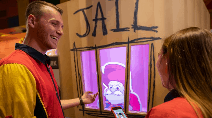 Guests share a laugh near Santa behind bars, Toy Story Land’s Stinky Pete style, ready for fun in the Play Disney Parks App!.