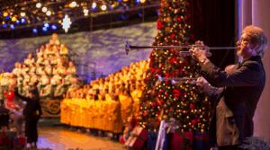 A trumpeter performs on stage beside a festive Christmas tree and robed choir during Epcot's Candlelight Processional.