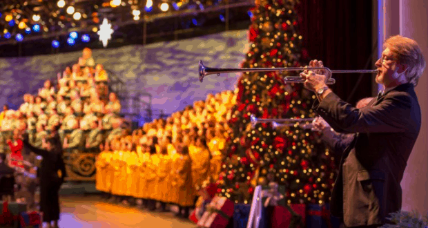 A trumpeter performs on stage beside a festive Christmas tree and robed choir during Epcot's Candlelight Processional.